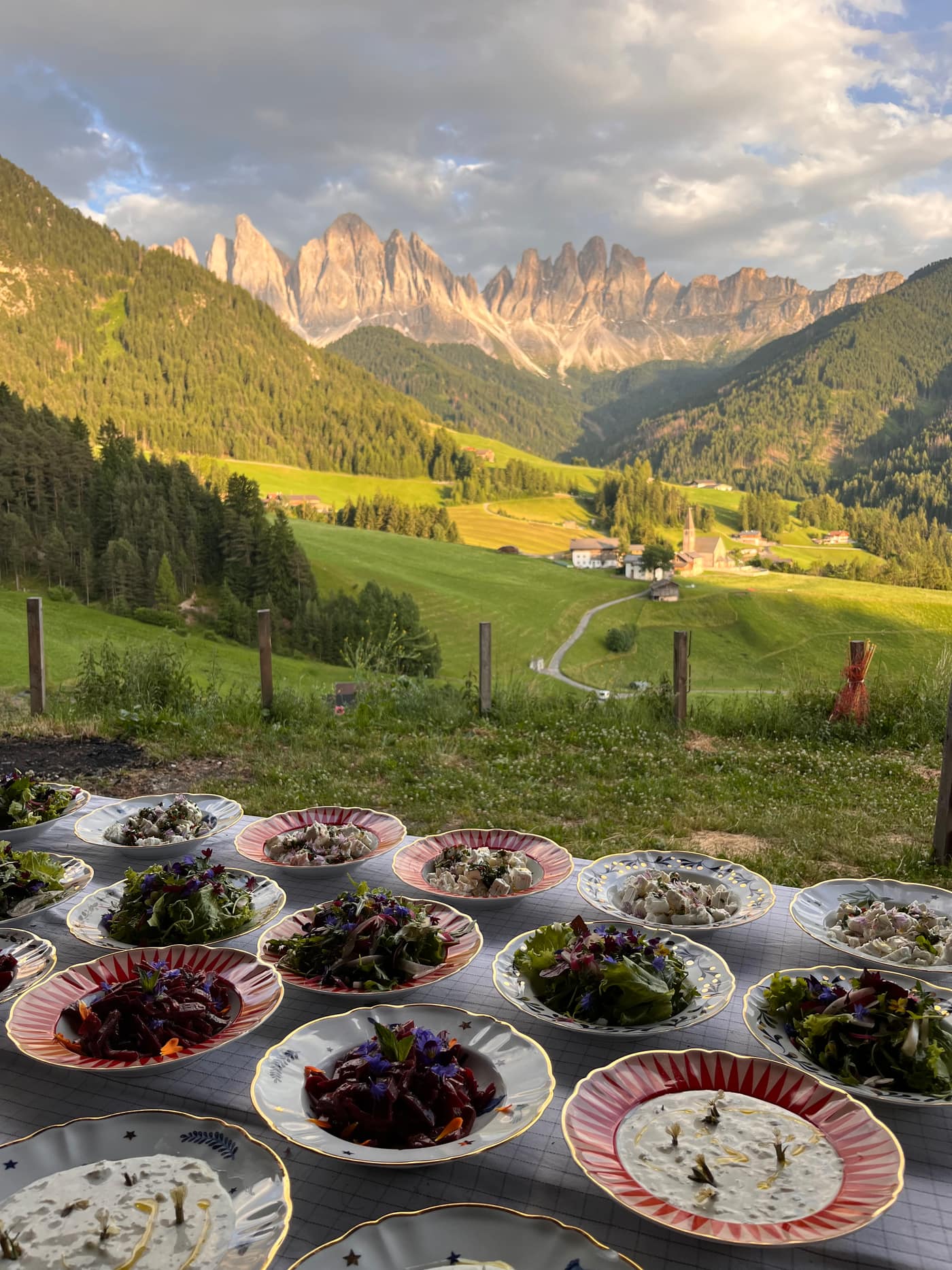 Langer Brunch-Tisch mit Tellern voller essbarer Blumen vor den Geislerspitzen in goldenem Licht